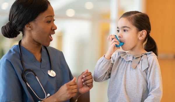 An ethnic girl with asthma is at a doctor's appointment. She is seated next to her African American doctor. The child is using a puffer. The doctor is watching and advising how to use the device properly.